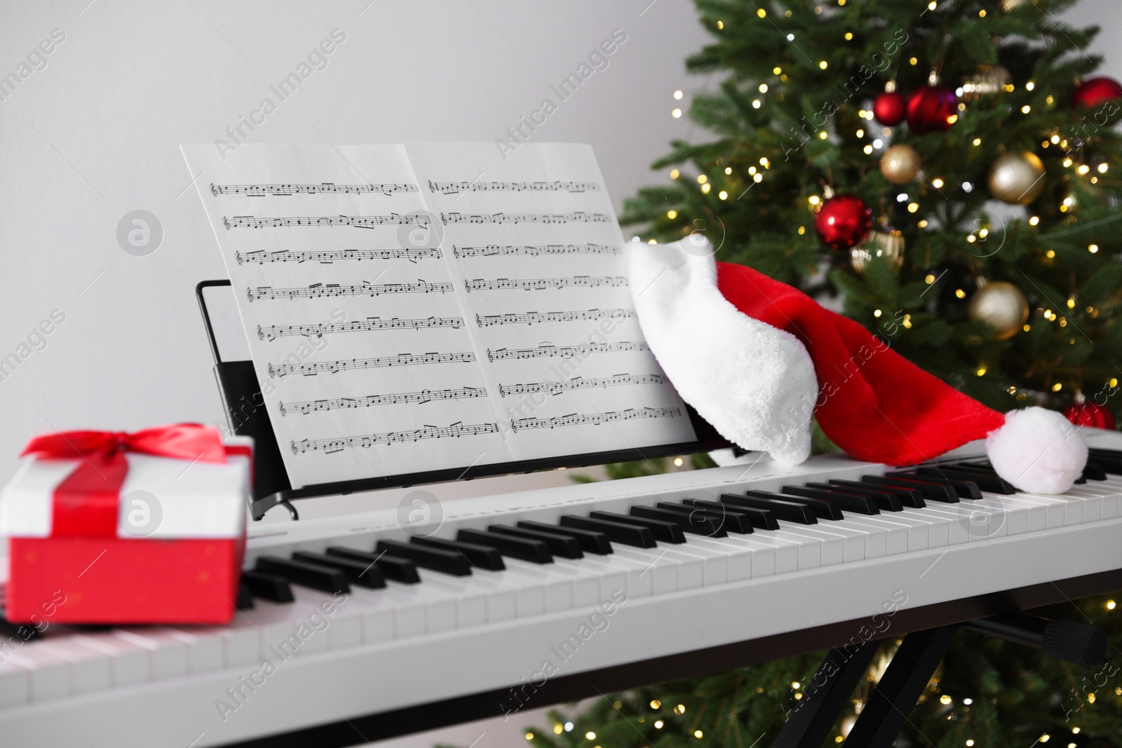 Synthesizer with music sheets, gift box and Santa hat near Christmas tree indoors, closeup Photo of Synthesizer with music sheets, gift box and Santa hat near Christmas tree indoors, closeup