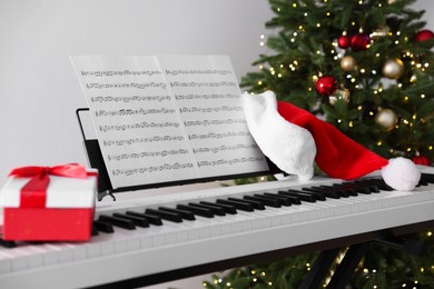 Photo of Synthesizer with music sheets, gift box and Santa hat near Christmas tree indoors, closeup