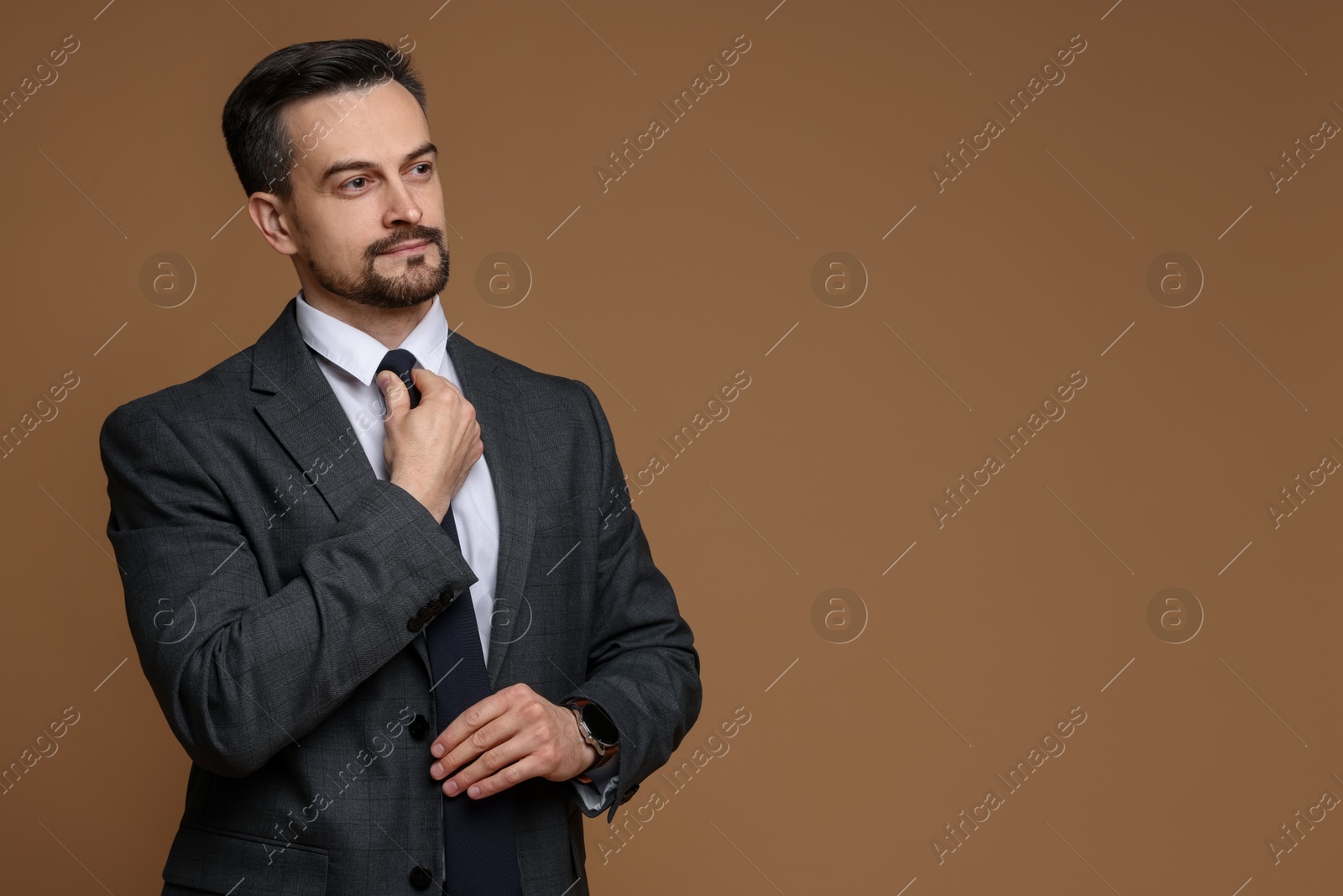 Photo of Confident man in classic suit straightening tie on brown background. Space for text