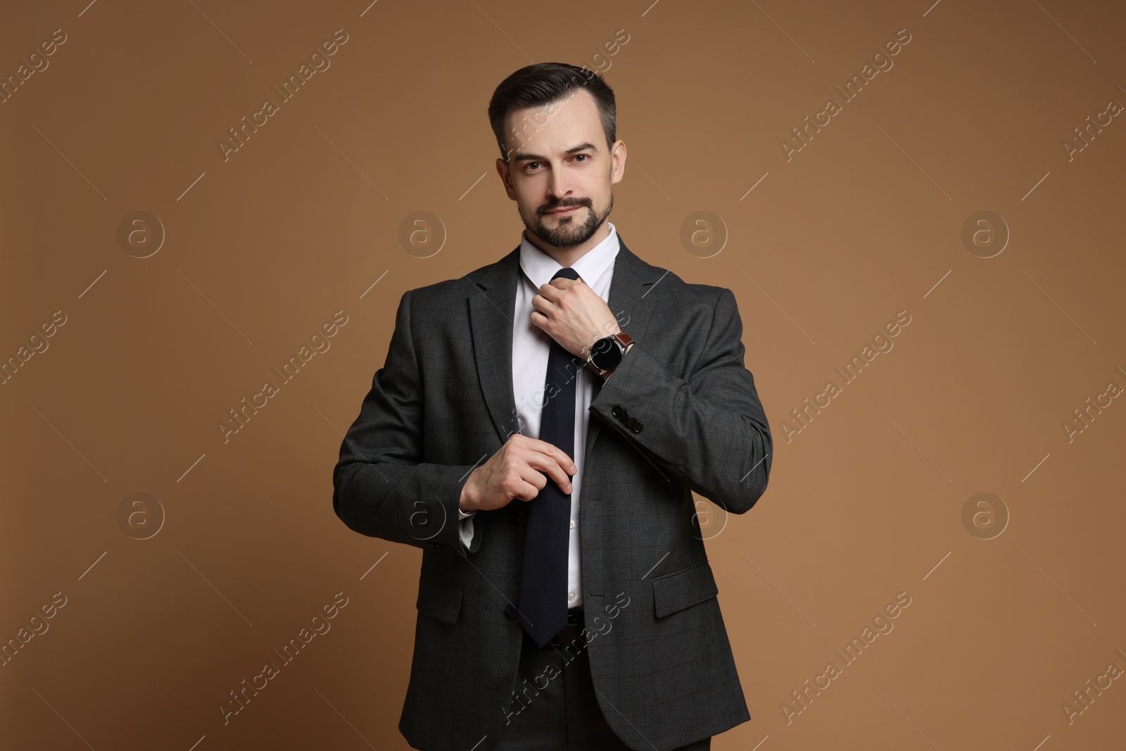 Photo of Confident man in classic suit straightening tie on brown background