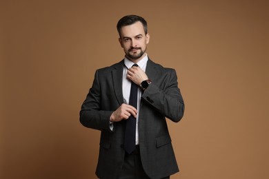 Confident man in classic suit straightening tie on brown background