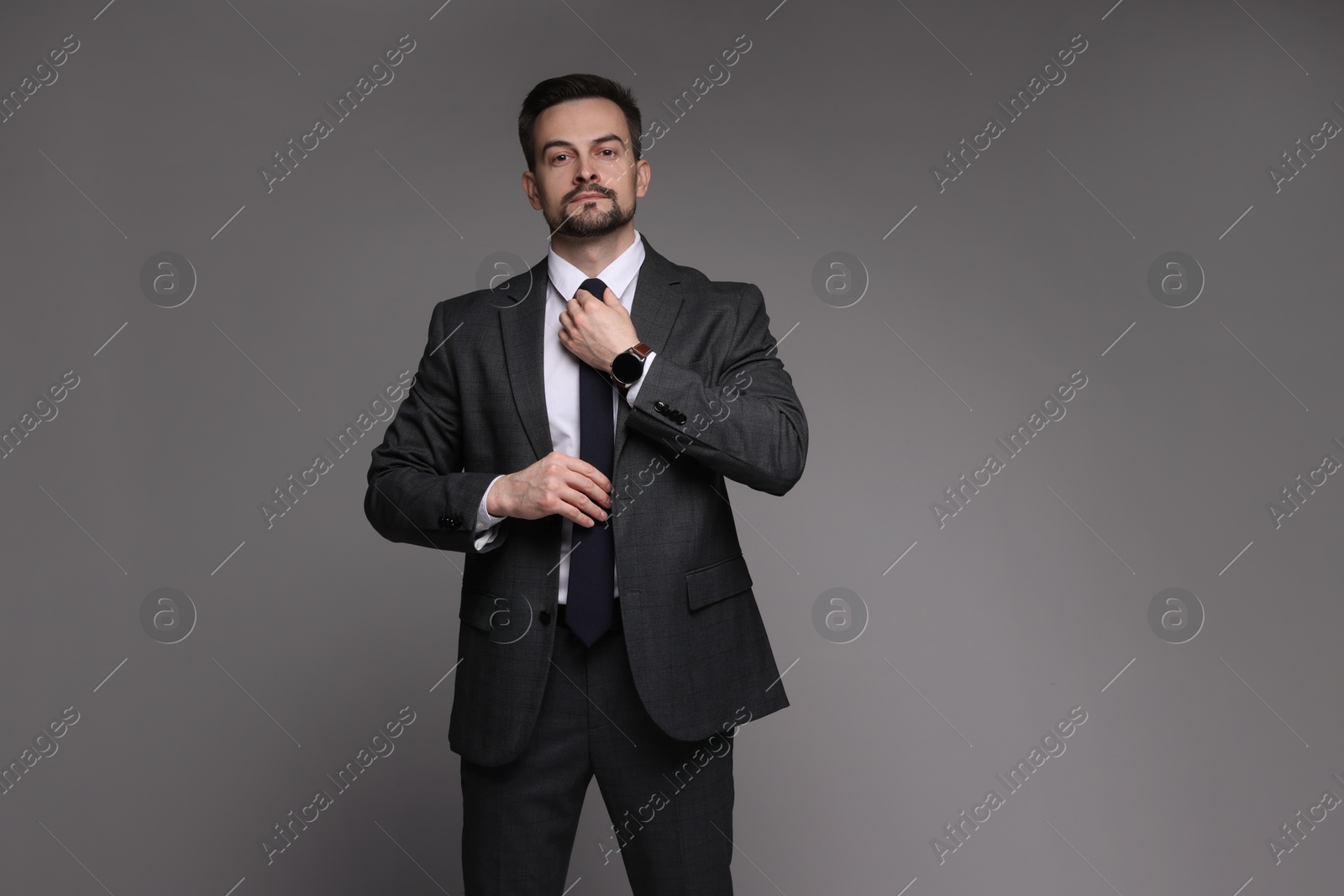 Photo of Confident man in classic suit straightening tie on grey background