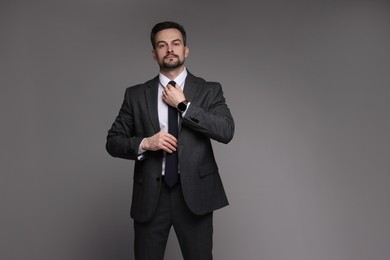 Confident man in classic suit straightening tie on grey background