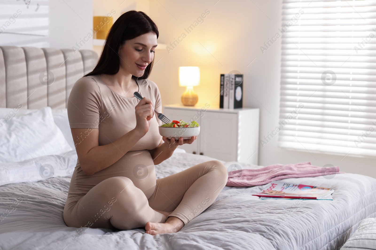 Photo of Beautiful pregnant woman eating vegetable salad on bed at home