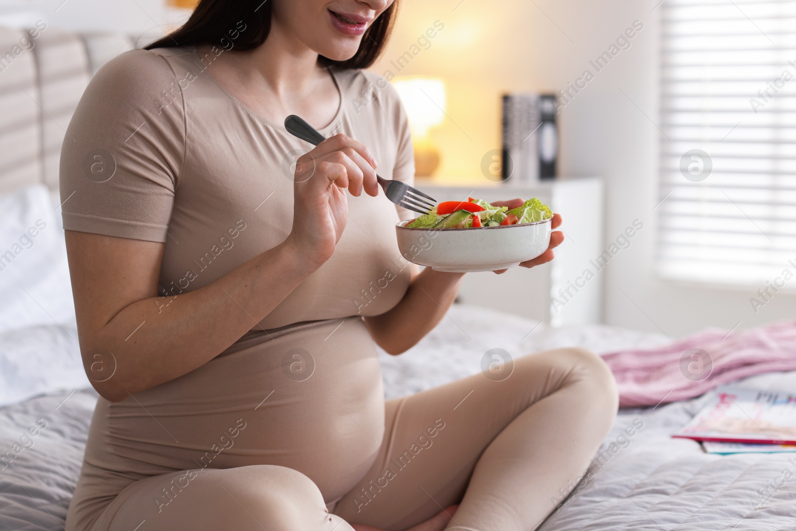 Photo of Pregnant woman eating vegetable salad on bed at home, closeup