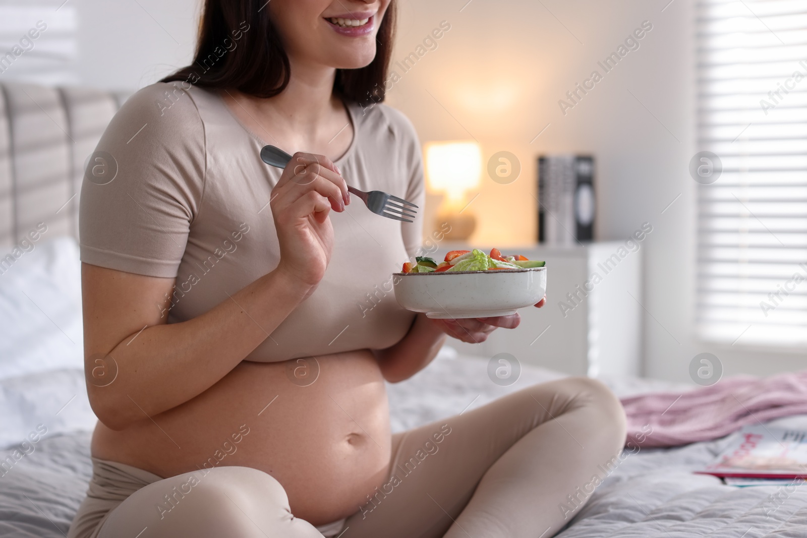 Photo of Pregnant woman eating vegetable salad on bed at home, closeup