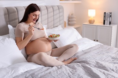 Photo of Beautiful pregnant woman eating vegetable salad on bed at home