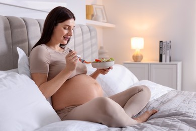 Photo of Beautiful pregnant woman eating vegetable salad on bed at home