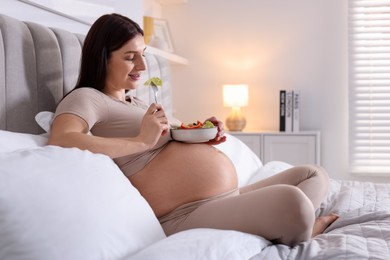 Photo of Beautiful pregnant woman eating vegetable salad on bed at home