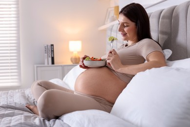 Beautiful pregnant woman eating vegetable salad on bed at home