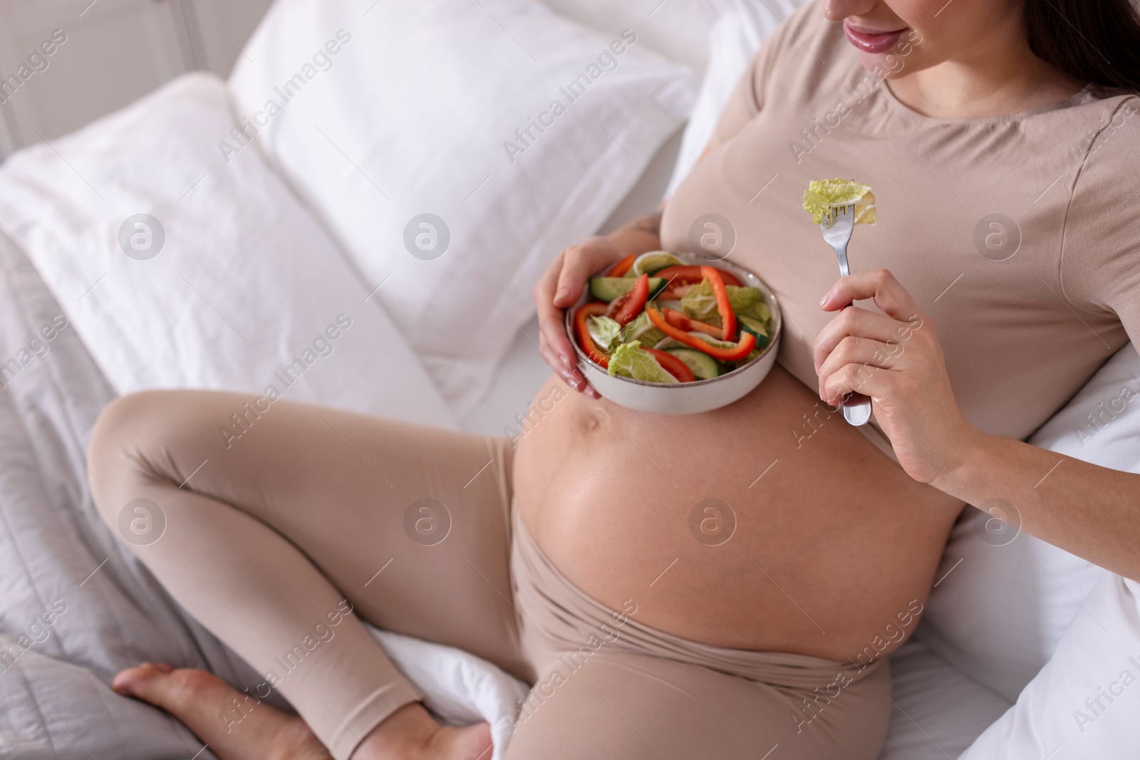 Photo of Pregnant woman eating vegetable salad on bed at home, closeup