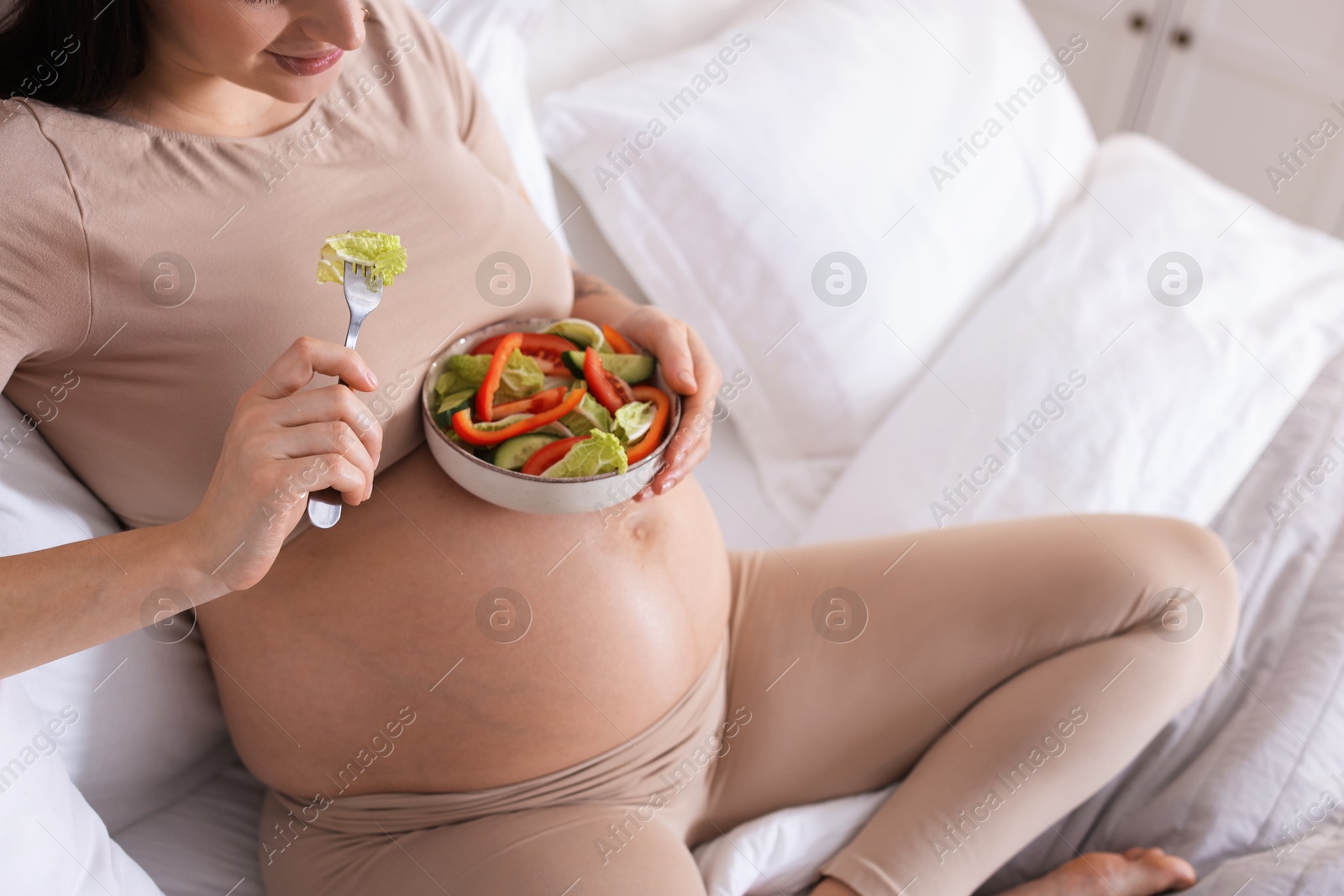 Photo of Pregnant woman eating vegetable salad on bed at home, closeup