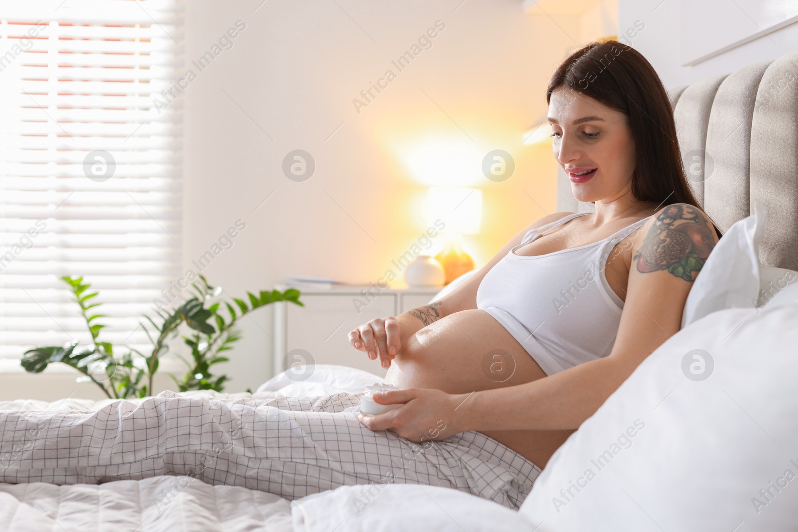 Photo of Pregnant woman with jar of cream on bed at home