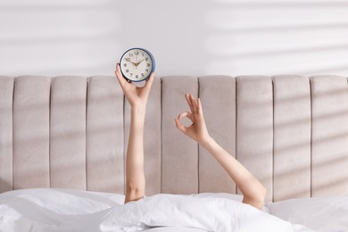 Woman with alarm clock showing ok gesture in bed at home, closeup. Good morning