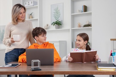 Photo of Smiling mother helping her children with homework at table indoors