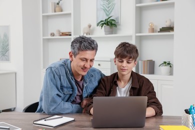 Photo of Smiling father and his son doing homework with laptop at table indoors