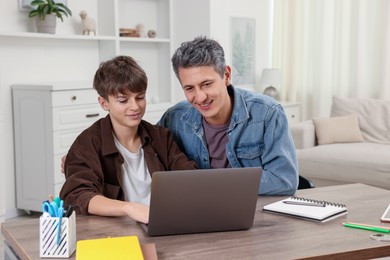 Photo of Smiling father and his son doing homework with laptop at table indoors