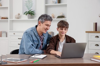 Photo of Smiling father and his son doing homework with laptop at table indoors