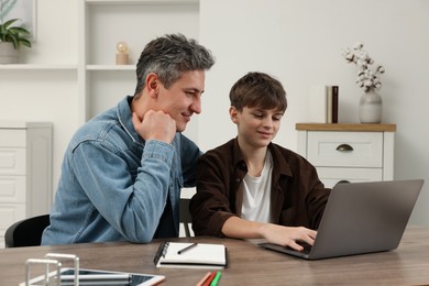 Photo of Smiling father and his son doing homework with laptop at table indoors