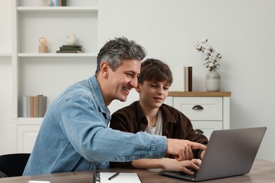 Photo of Smiling father and his son doing homework with laptop at table indoors