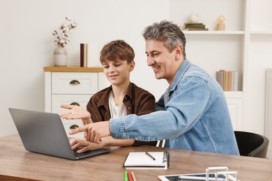 Smiling father and his son doing homework with laptop at table indoors