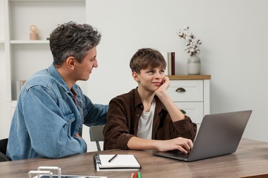 Photo of Father and his tired son doing homework with laptop at table indoors