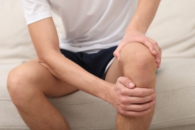 Photo of Young man suffering from pain in knee on sofa at home, closeup