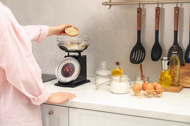 Photo of Woman adding peanuts into bowl on mechanical kitchen scale at white marble countertop indoors, closeup