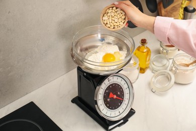 Photo of Woman adding peanuts into bowl on mechanical kitchen scale at white marble countertop indoors, closeup