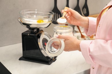 Photo of Woman holding spoon with flour near mechanical kitchen scale at white marble countertop indoors, closeup