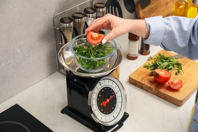 Photo of Woman adding tomato into bowl on mechanical kitchen scale at white marble countertop indoors, closeup