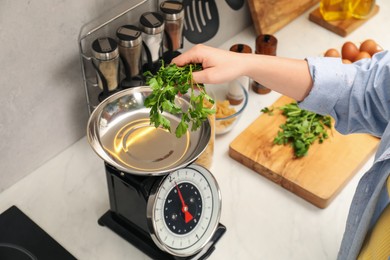 Photo of Woman putting parsley into bowl on mechanical kitchen scale at white marble countertop indoors, closeup