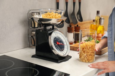 Photo of Woman holding container of raw pasta near mechanical kitchen scale at white marble countertop indoors, closeup