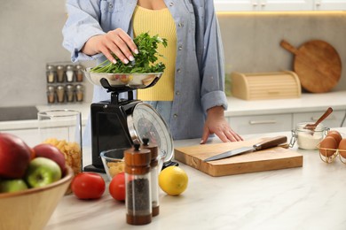 Photo of Woman putting parsley into bowl on mechanical kitchen scale at white marble table indoors, closeup