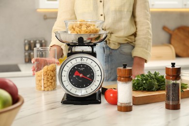 Photo of Woman holding container of raw pasta near mechanical kitchen scale at white marble countertop indoors, closeup