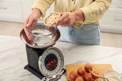 Photo of Woman adding peanuts into bowl on mechanical kitchen scale at white marble table indoors, closeup