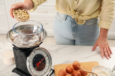 Photo of Woman adding peanuts into bowl on mechanical kitchen scale at white marble table indoors, closeup