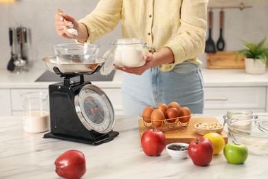 Photo of Woman adding flour into bowl on mechanical kitchen scale at white marble table indoors, closeup