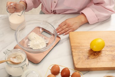 Photo of Woman with measuring cup of milk near electronic kitchen scale at white marble table indoors, closeup