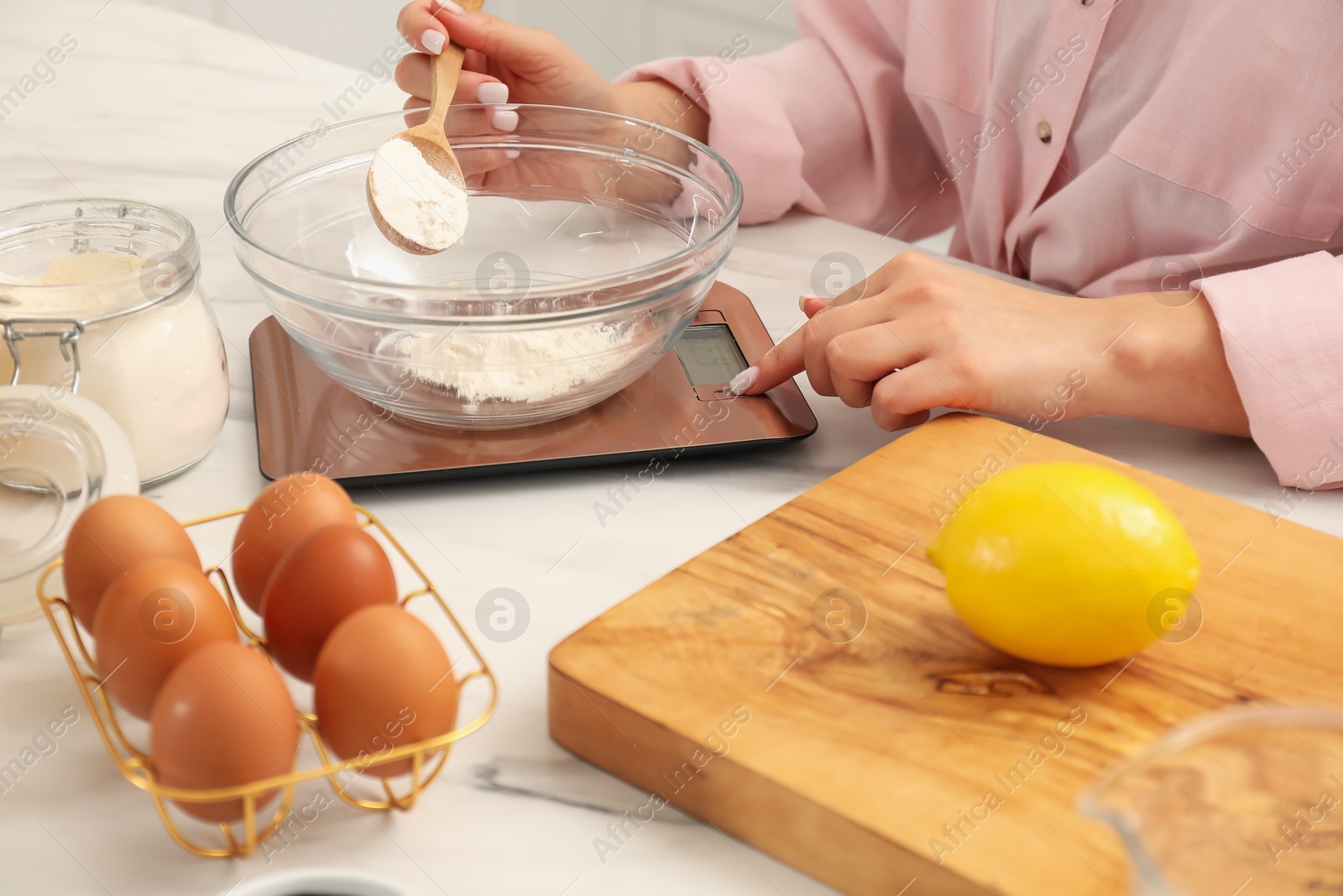 Photo of Woman adding flour into bowl on electronic kitchen scale at white marble table indoors, closeup