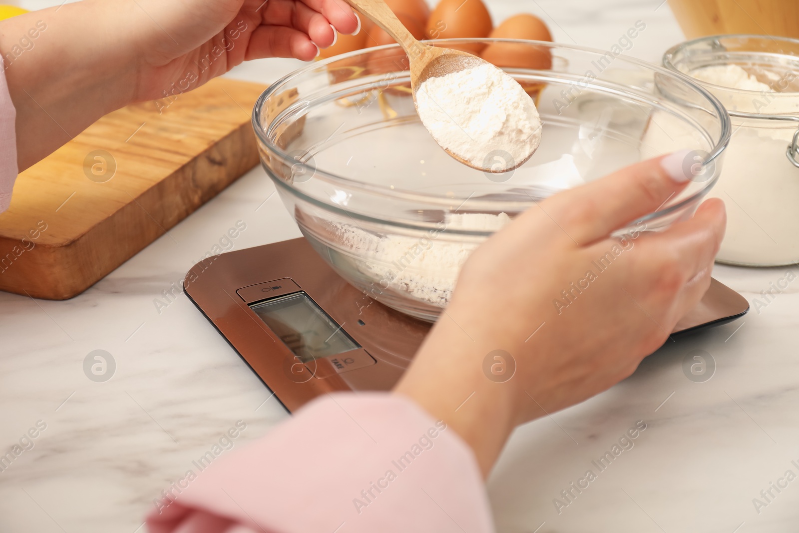 Photo of Woman adding flour into bowl on electronic kitchen scale at white marble table indoors, closeup