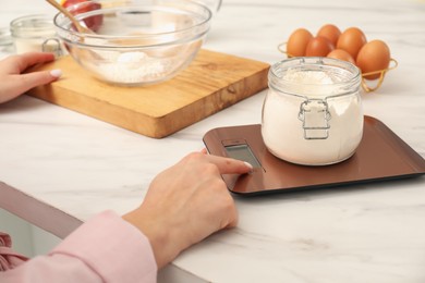 Photo of Woman weighing jar of flour on electronic kitchen scale indoors, closeup