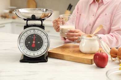 Photo of Woman with measuring cup of milk near mechanical kitchen scale at white marble table indoors, closeup