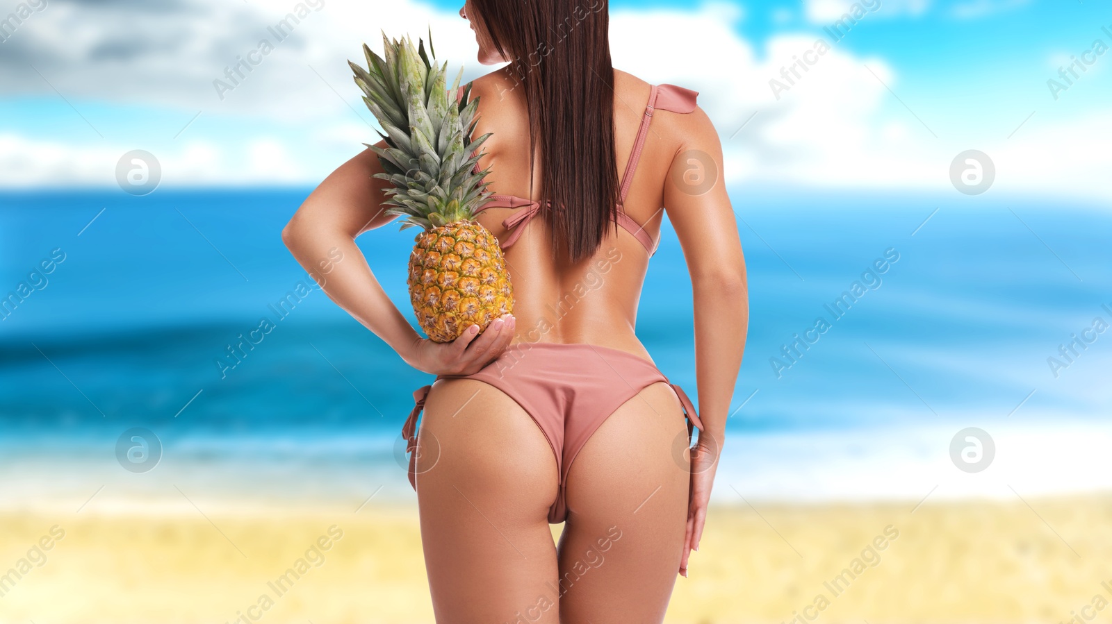 Image of Sexy young woman in pink swimsuit holding pineapple on sandy beach near blue sea, closeup. Back view. Banner design