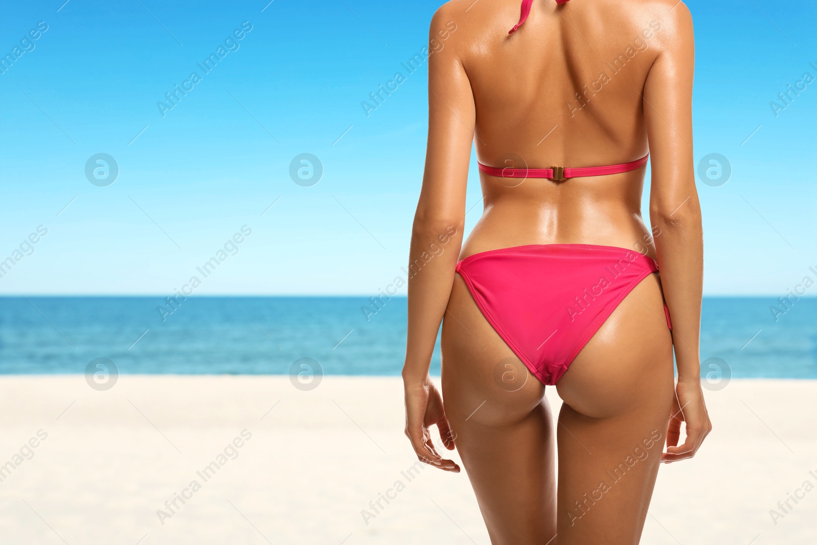 Image of Sexy young woman in pink swimsuit on sandy beach near blue sea, closeup. Girl has perfect tanned body, back view. Space for text