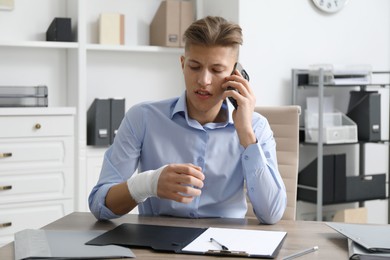 Man with wrist wrapped in medical bandage talking on smartphone at table indoors