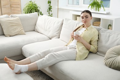 Woman with cup of drink and medical bandage on her foot at home