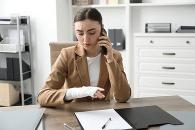 Photo of Woman with wrist wrapped in medical bandage talking on smartphone at table indoors