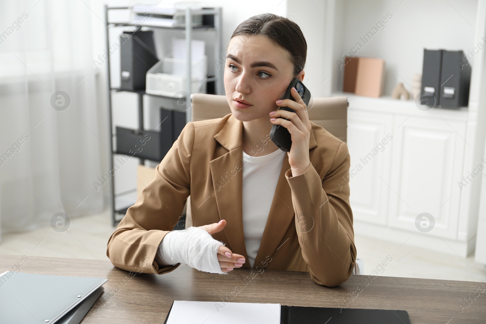 Photo of Woman with wrist wrapped in medical bandage talking on smartphone at table indoors