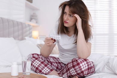 Photo of Unhappy woman with blister of pills suffering from hangover on bed at home